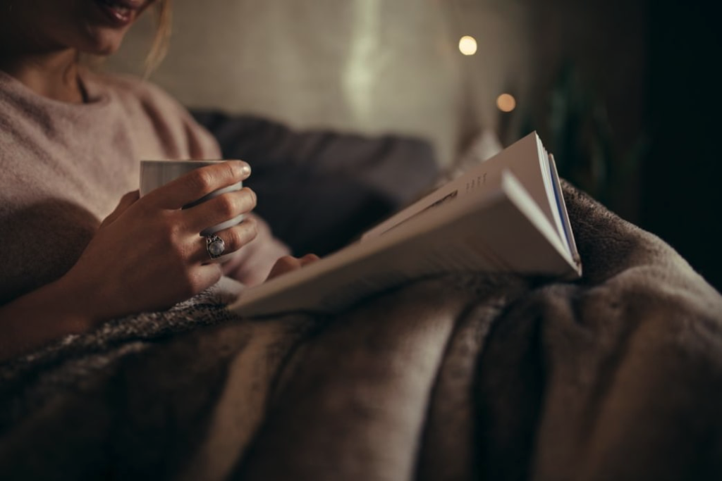 A woman in a cozy blanket and cup in her hand, reading a book.