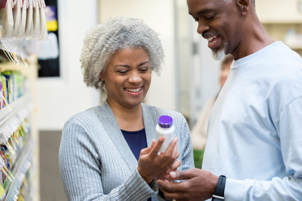 Senior adult African American man and woman are reading labels on bottle of medication or vitamins while shopping together in supermarket or pharmacy. Represents vitamin D supplements. 