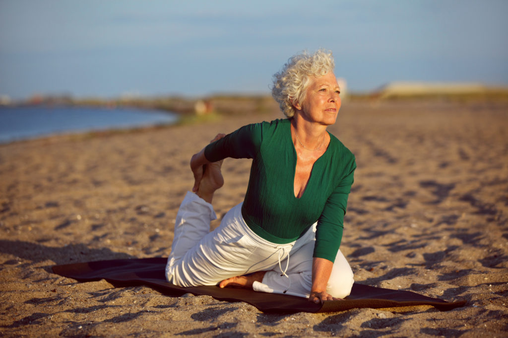 Senior woman in stretching position by the sea at morning. Elderly woman doing yoga on the beach.