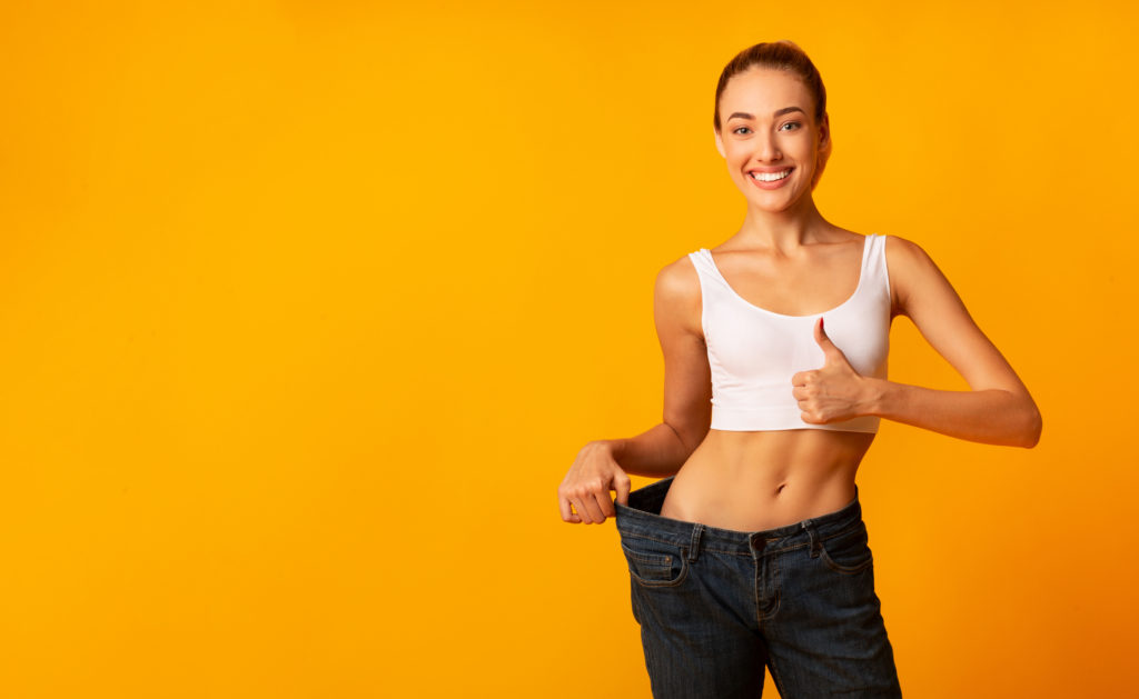 Weight Loss. Girl In Oversize Jeans Gesturing Thumbs Up Smiling At Camera Standing On Yellow Studio Background. Copy Space