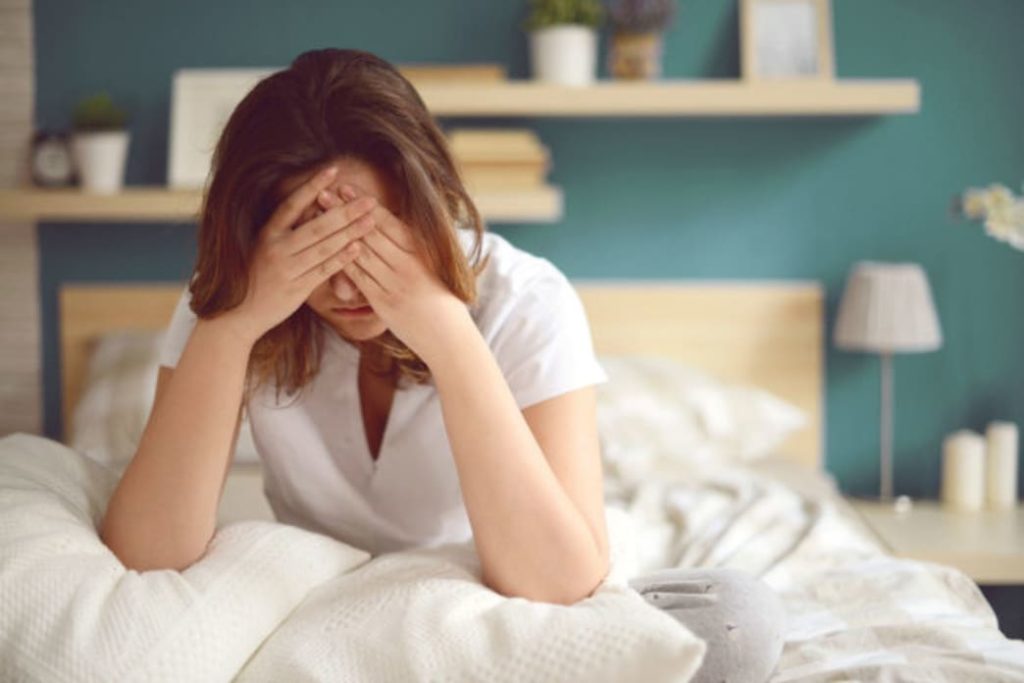 A woman sitting up in bed and holding her head with worry. 