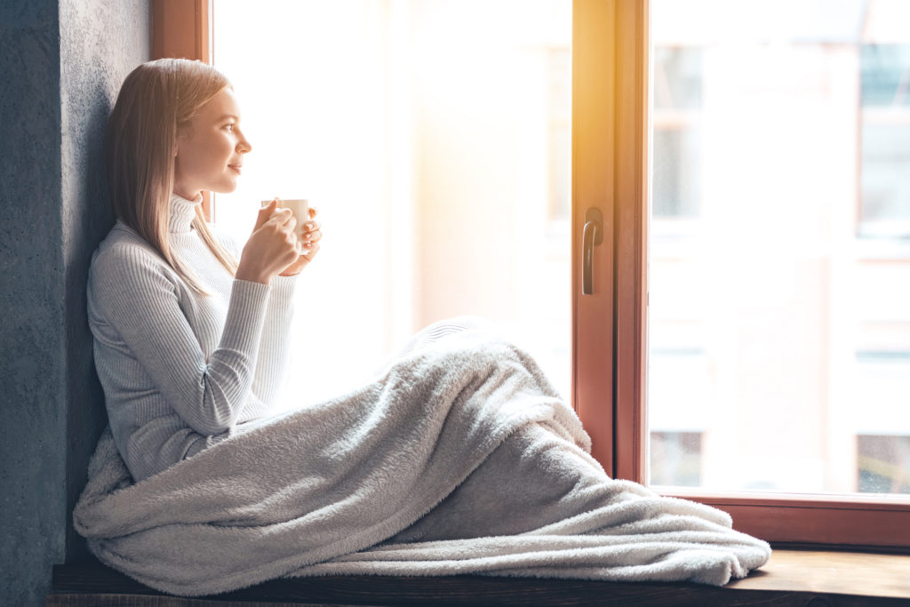 Side view of beautiful young woman holding coffee cup and looking through window while sitting at windowsill at home