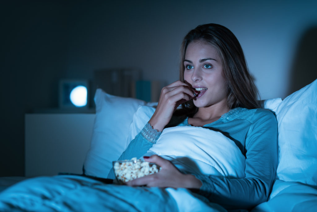 Happy woman relaxing in bed and watching movies on tv late at night, she is eating popcorn