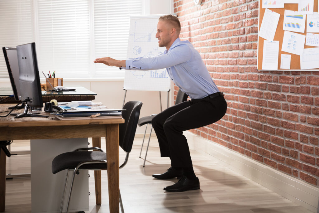Businessman Doing Exercise In Office shows standing behind the chair and arms extended