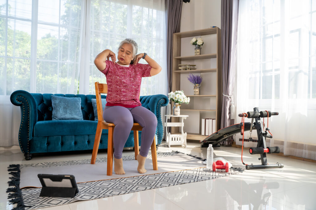 Senior woman sitting on a wooden chair and slowly rotate both shoulders, doing with training exercise online on tablet, workout online from home concept