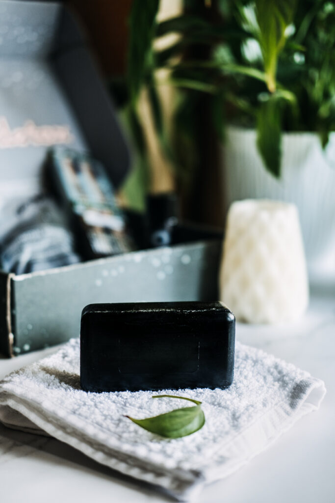 Lifestyle still life with black charcoal soap on beauty box background. Handmade soap with activated carbon on cotton towel and marble gray background